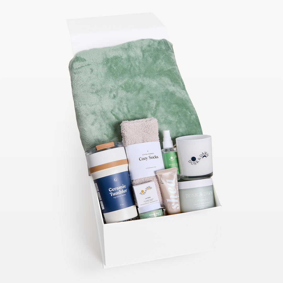 Comfort & Care Sage Green, white gift box, lid open. Sage faux-fur throw, cream Cozy Socks, Ceramic Tumbler, spray, cream candle, Shea bar, cucumber tin. Giften Market.