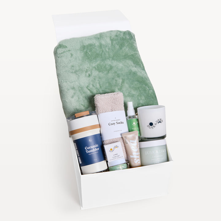 Comfort &amp; Care Sage Green, white gift box, lid open. Sage faux-fur throw, cream Cozy Socks, Ceramic Tumbler, spray, cream candle, Shea bar, cucumber tin. Giften Market.