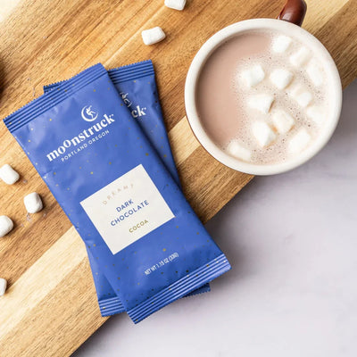 Two Moonstruck Dreamy Dark Chocolate Hot Cocoa single-serve pouches beside a mug of cocoa with marshmallows
