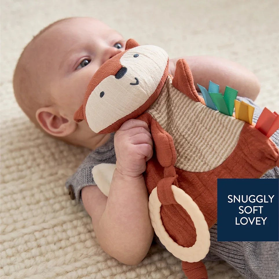 Baby holding fox-shaped sensory toy with teether and colorful tags on soft rug