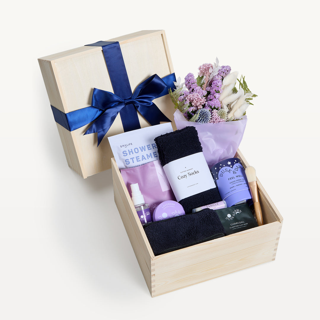 Grace & Gratitude Lavender crate open in front, bow-tied lid behind. Pink and lavender dried bouquet, black Cozy Socks, SpaLife shower steamer, Feel Well tea, lavender spray, charcoal clay mask, wooden spoon. Giften Market.