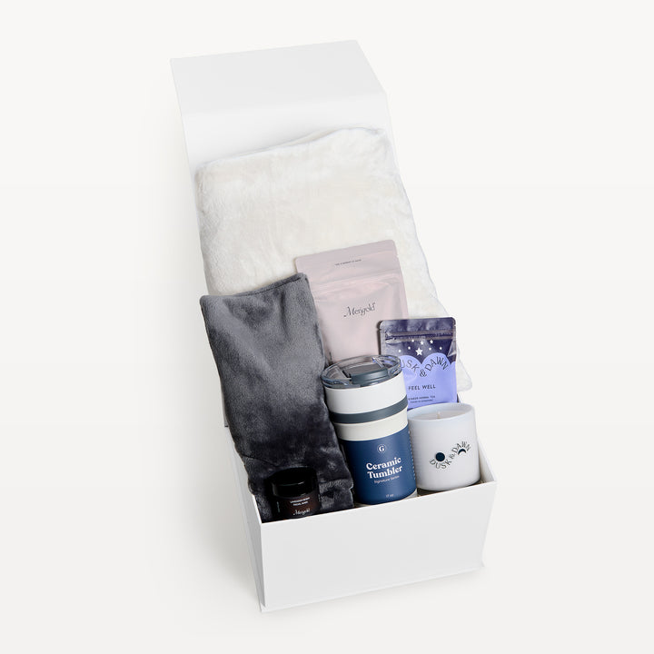 Healing Harmony Cream, white gift box, lid open. Cream faux-fur throw over a charcoal throw, Marigold pouches, Feel Well tea, Ceramic Tumbler, cream candle, small amber jar. Giften Market.