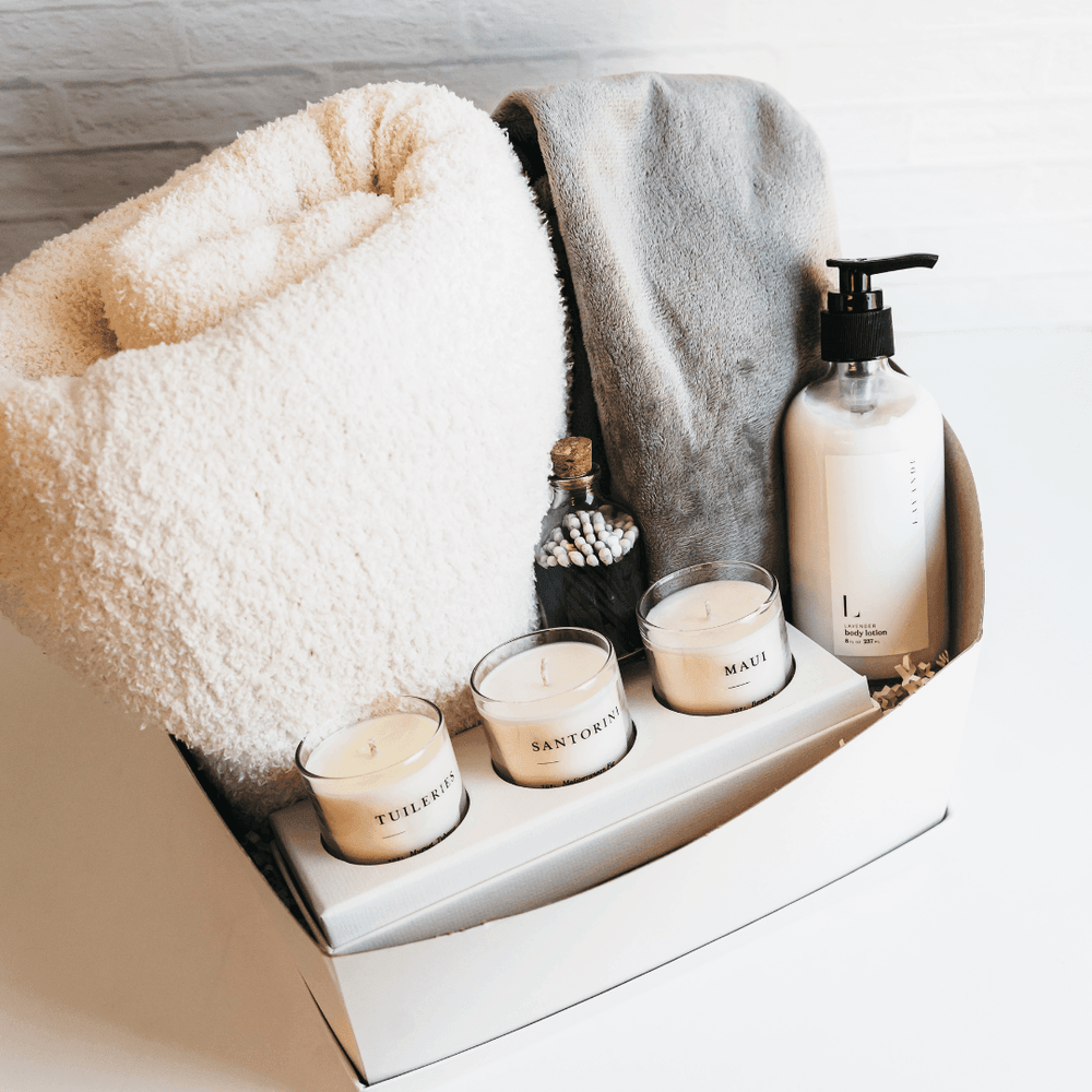 White gift box with candles, plush blankets, body lotion, and matches arranged neatly