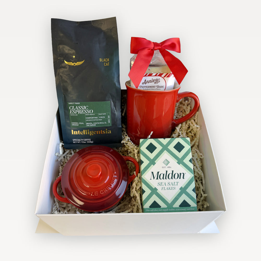 The Holiday Kitchen Espresso Edition gift box in Cerise: Le Creuset Vancouver mug and mini cocotte in red, Intelligentsia Black Cat espresso, Maldon sea salt flakes, and Annie B's peppermint bark with a red bow, in a white gift box with ivory crinkle