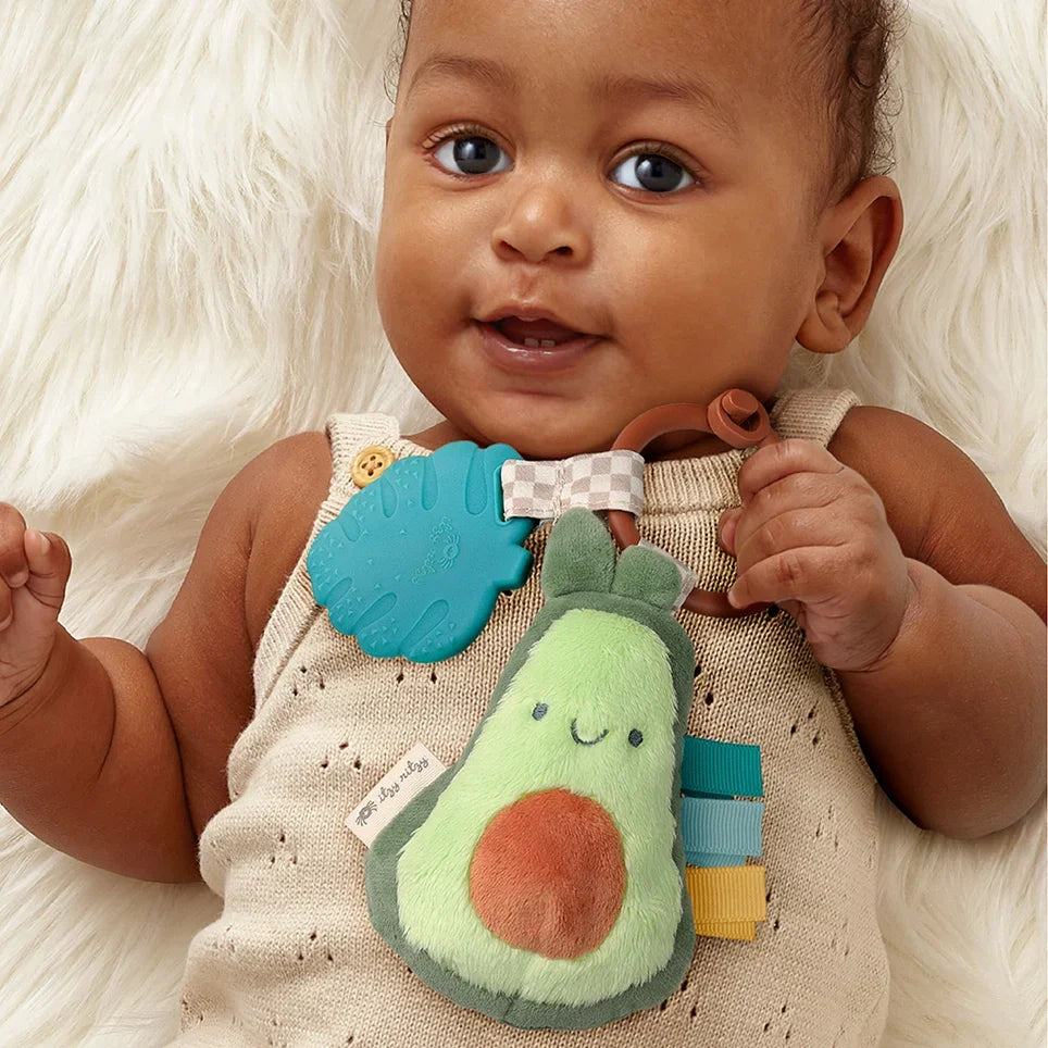 Smiling baby holding avocado plush teether toy and blue silicone teething leaf