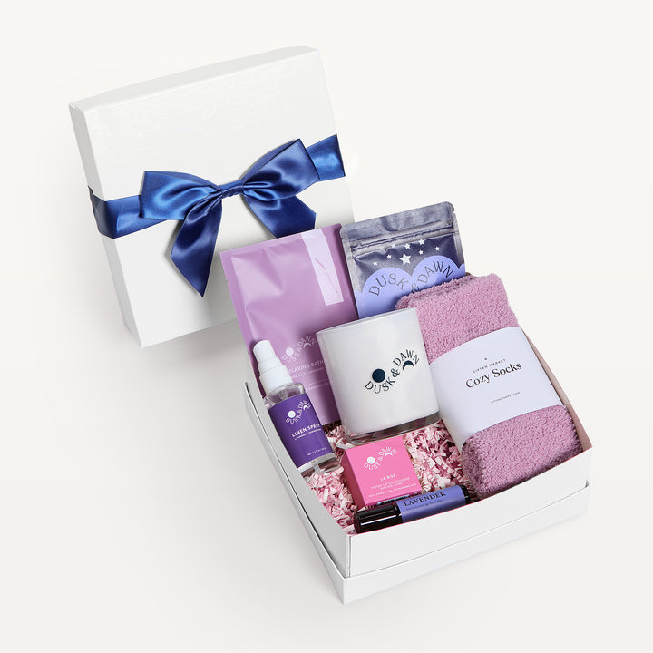 Lavender Luxuries, white shallow box open in front with the bow-tied lid propped behind. Lavender pouches, pink Cozy Socks, cream candle, lavender spray, lip balm, Dusk &amp; Dawn tin, pink crinkle. Giften Market.