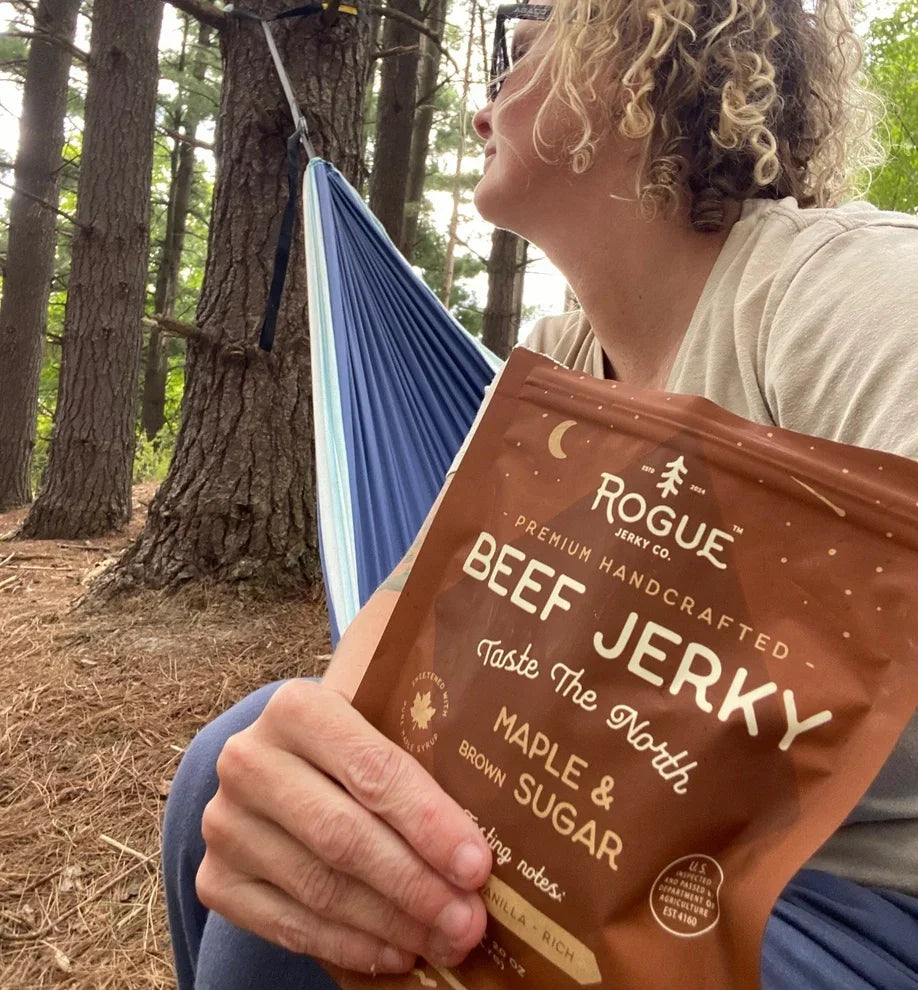 Person holding maple and brown sugar beef jerky outdoors by a hammock in the woods