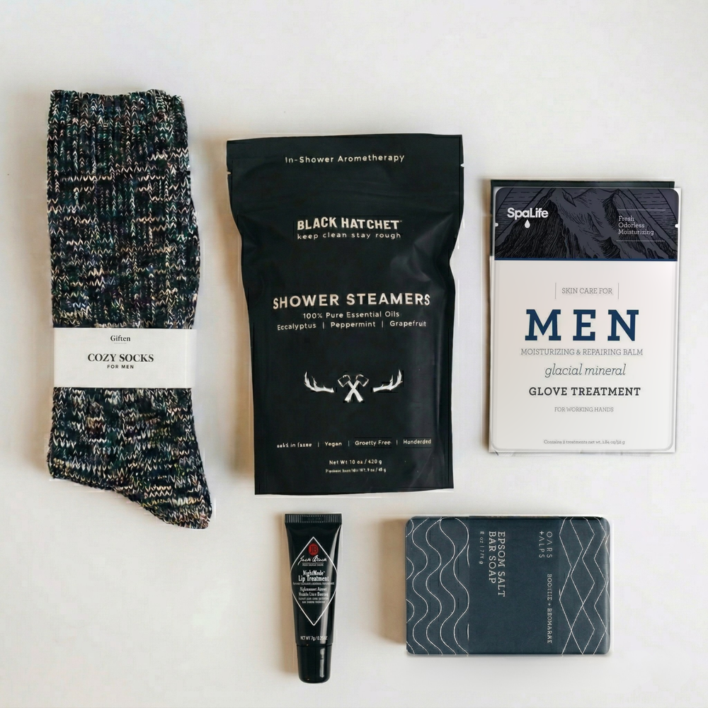 Flat lay of the men's self-care gift box contents: cozy socks, shower steamers, glacial mineral hand glove treatment, lip treatment and epsom salt bar soap