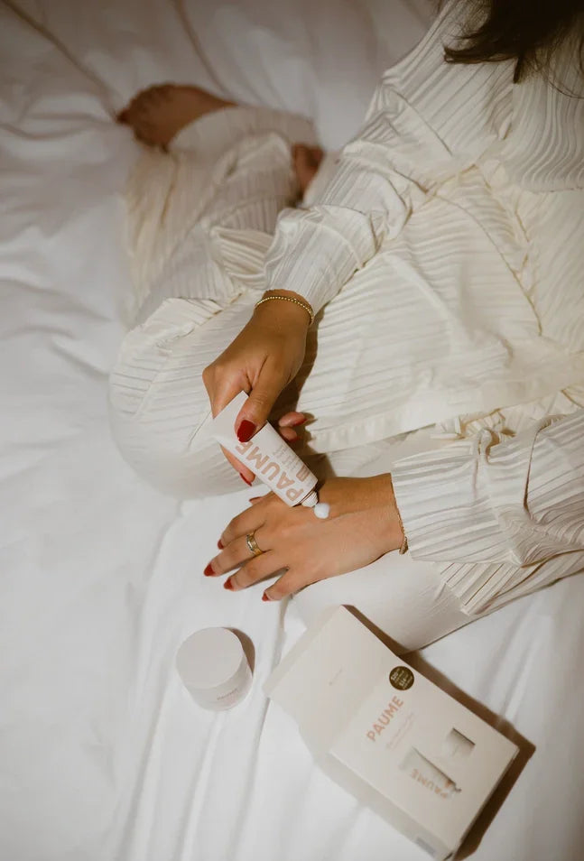 Woman applying hand cream from Paume gift set on white bedding, with boxed set and jar