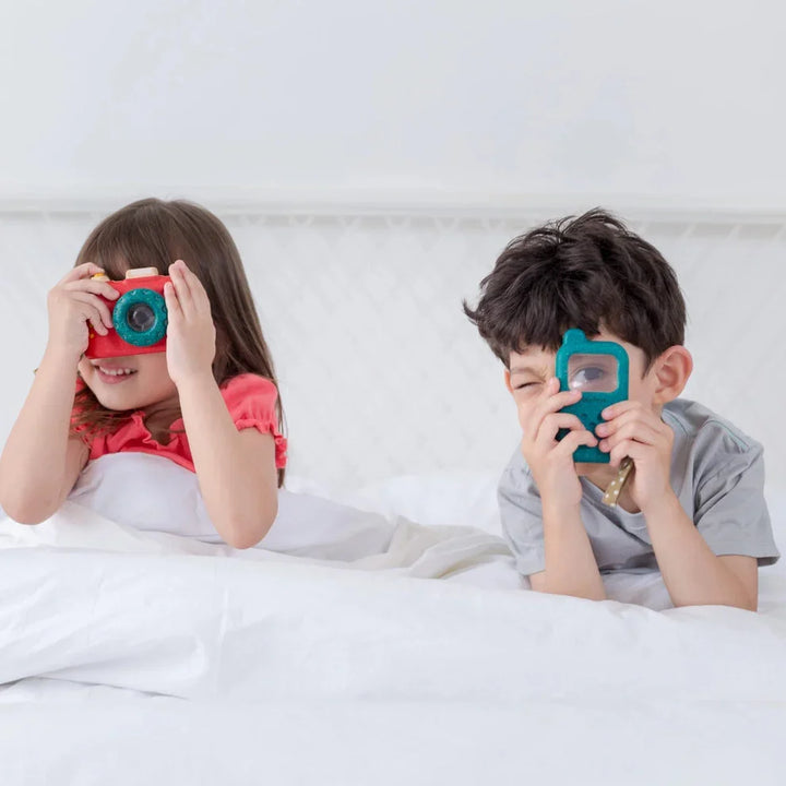Two children on a bed playing with colorful toy camera and phone, white bedding background