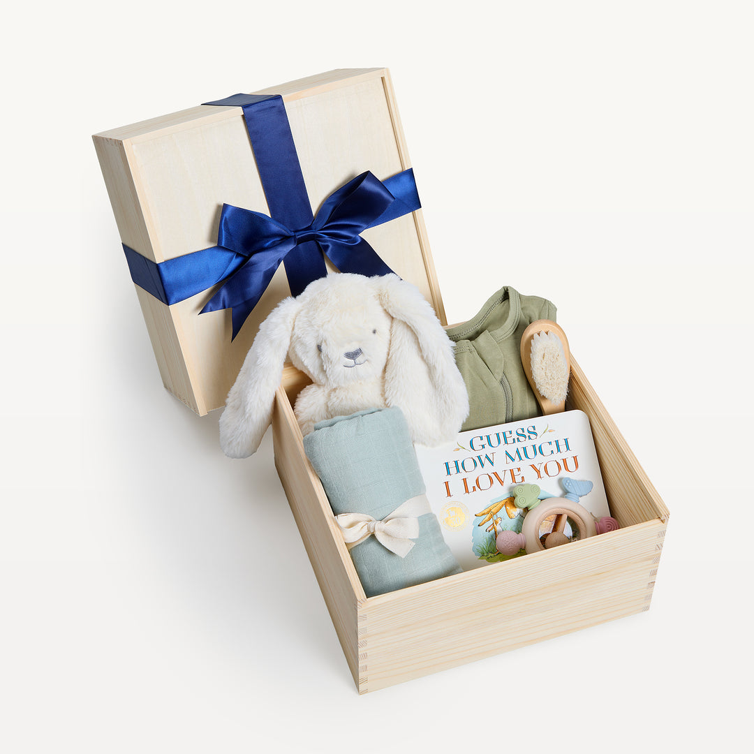 Nest & Nurture crate open in front, bow-tied lid behind. Cream plush bunny, sage onesie and swaddle, Guess How Much I Love You board book, wooden brush and teether. Giften Market.