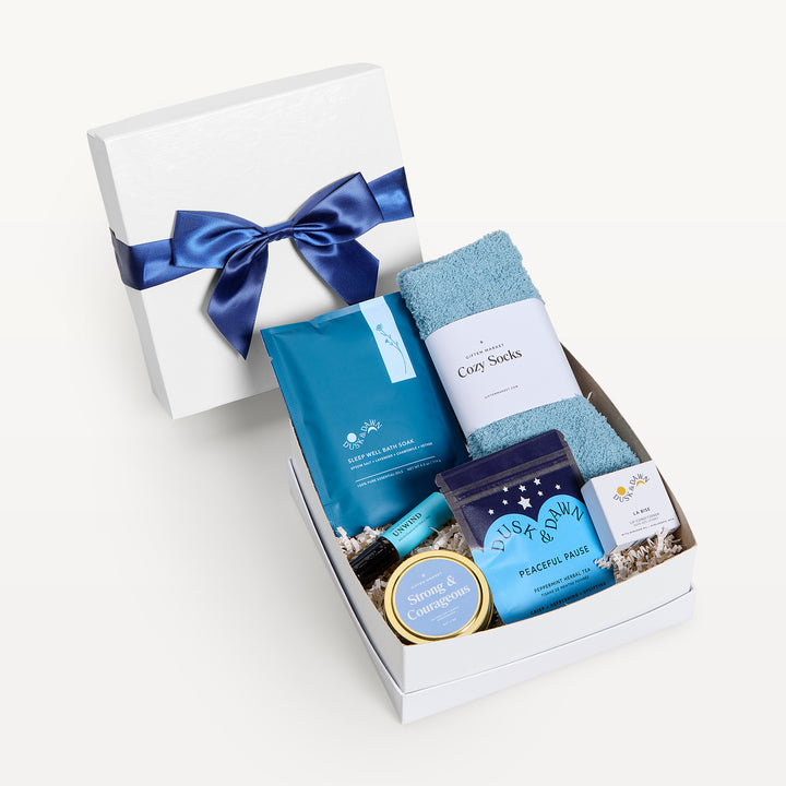 Peaceful Pause, white shallow box open in front, bow-tied lid behind. Teal Cozy Socks, Sleep Well bath soak pouch, Peaceful Pause tea, Strong &amp; Courageous tin, lip balm, small yellow box. Giften Market.