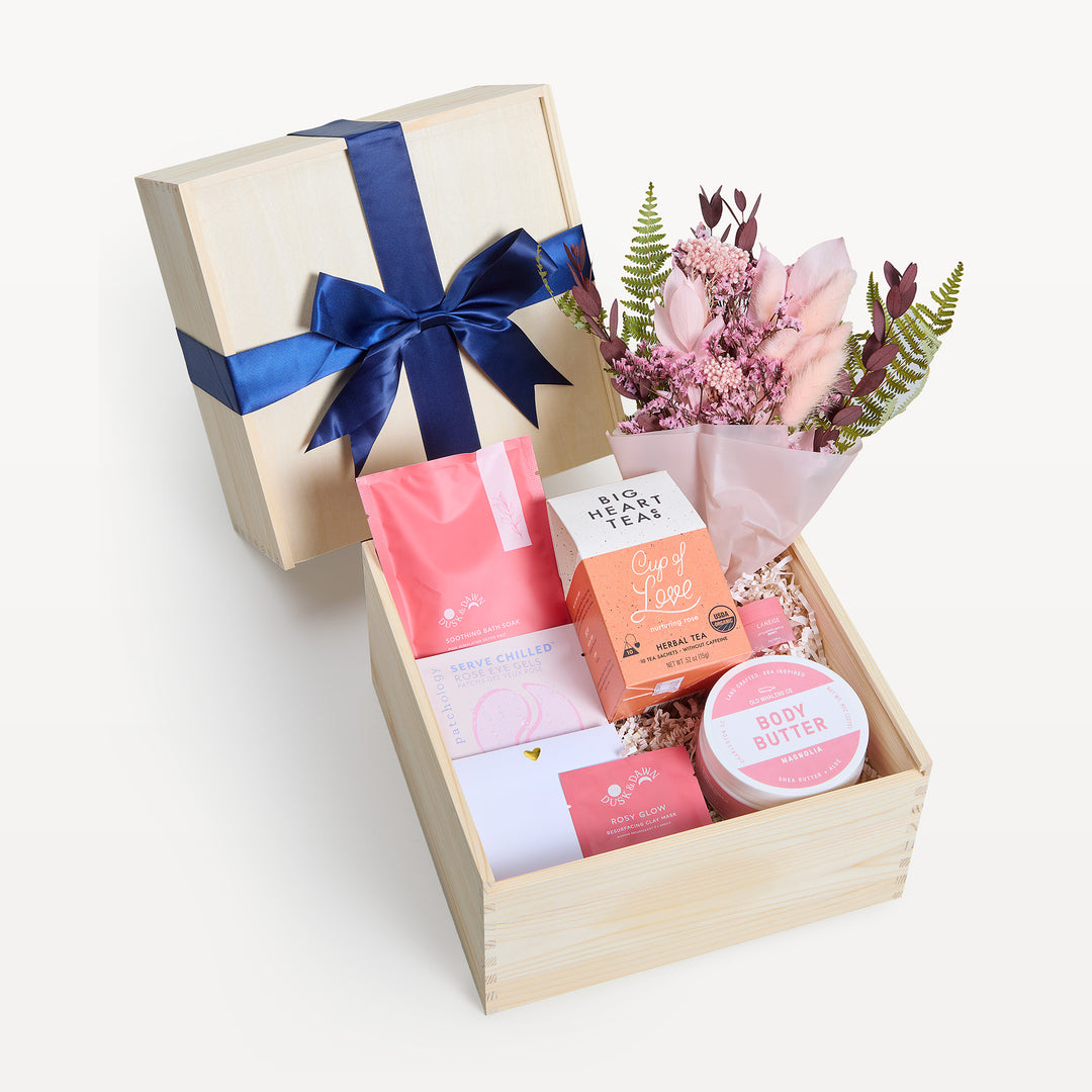 Radiant Relaxation crate open in front with the bow-tied lid propped behind. Pink dried-flower bouquet, Big Heart Teas Cup of Love, Rosy Glow face mask, body butter, crinkle paper. Giften Market.