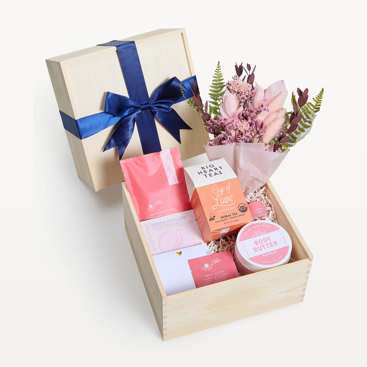 Radiant Relaxation crate open in front with the bow-tied lid propped behind. Pink dried-flower bouquet, Big Heart Teas Cup of Love, Rosy Glow face mask, body butter, crinkle paper. Giften Market.