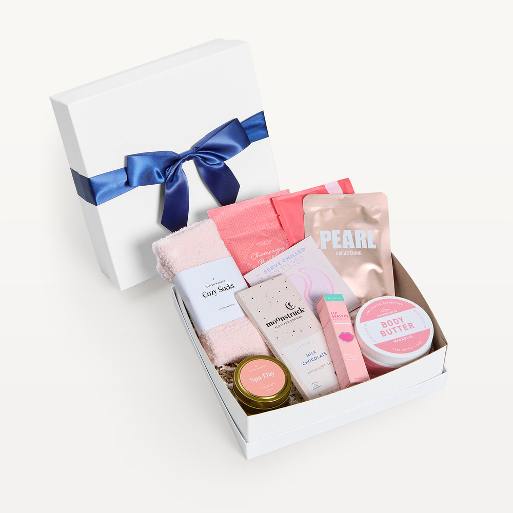 Rosy Outlook Deluxe, white shallow box open in front, bow-tied lid behind. Pink Cozy Socks, Pearl brightening mask, Patchology, Moonstruck milk chocolate, Grapefruit body butter, Spa Day candle tin, lip treatment. Giften Market.