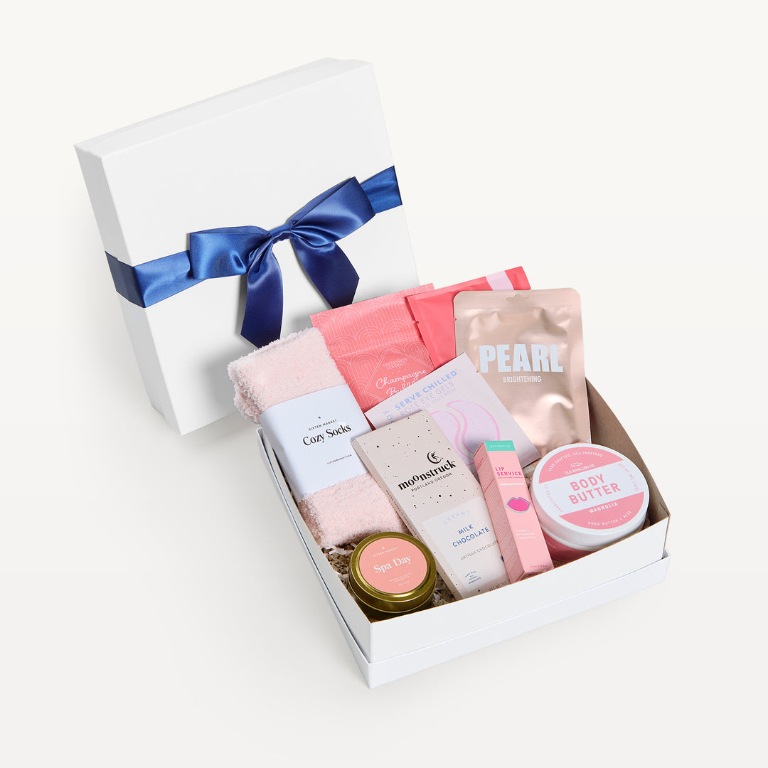 Rosy Outlook Deluxe, white shallow box open in front, bow-tied lid behind. Pink Cozy Socks, Pearl brightening mask, Patchology, Moonstruck milk chocolate, Grapefruit body butter, Spa Day candle tin, lip treatment. Giften Market.