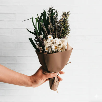 Hand holding the Savannah Dried Flower Mini Bouquet in taupe paper against white brick