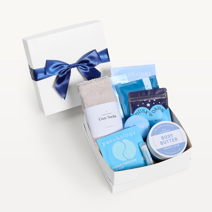 Seaside Escape Classic, white shallow box open in front, bow-tied lid behind. Cream Cozy Socks, Patchology Serve Chilled, blue pouches, Peaceful Pause tea, Coastal Calm body butter, lip balm. Giften Market.