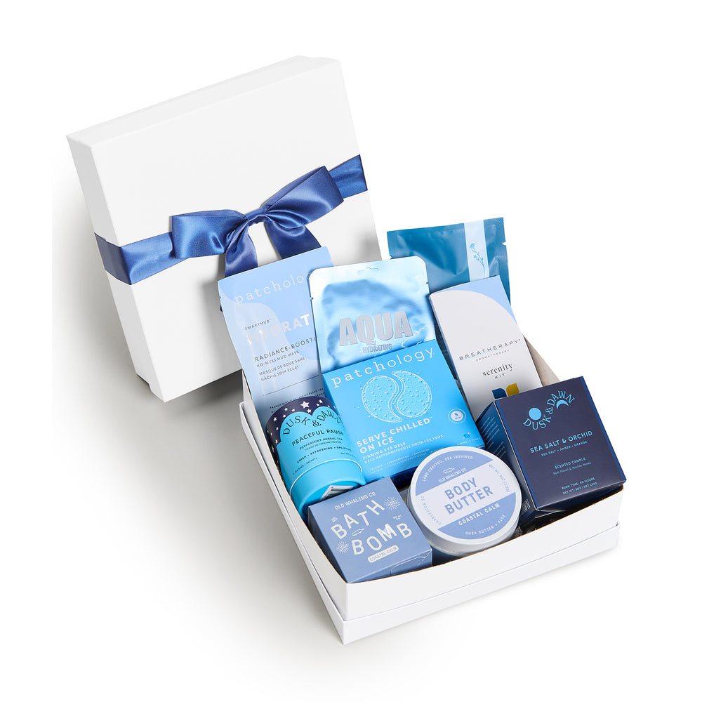 Seaside Escape Deluxe, white shallow box open in front, bow-tied lid behind. Patchology Radiance Boost and Serve Chilled, Aqua mask, Breatherapy Serenity, Peaceful Pause tea, Sea Salt & Orchid candle, Coastal Calm body butter, bath bomb. Giften Market.