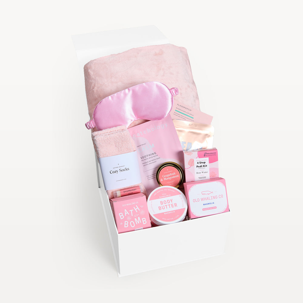 Self-Care Sunday, white gift box, lid open. Blush faux-fur throw, pink satin sleep mask, pink Cozy Socks, Patchology Soothing mask, Grapefruit & Mangosteen tin, Magnolia body butter, 4 Step Pedi Kit, Old Whaling bath bomb and soap. Giften Market.