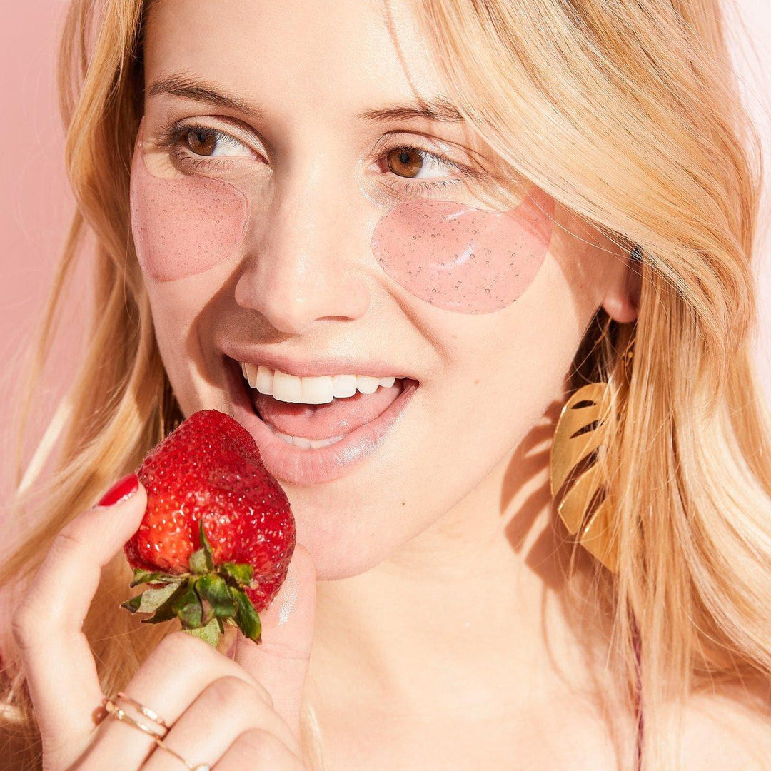 Woman wearing pink under eye gel patches, holding a fresh strawberry, gold leaf earring visible