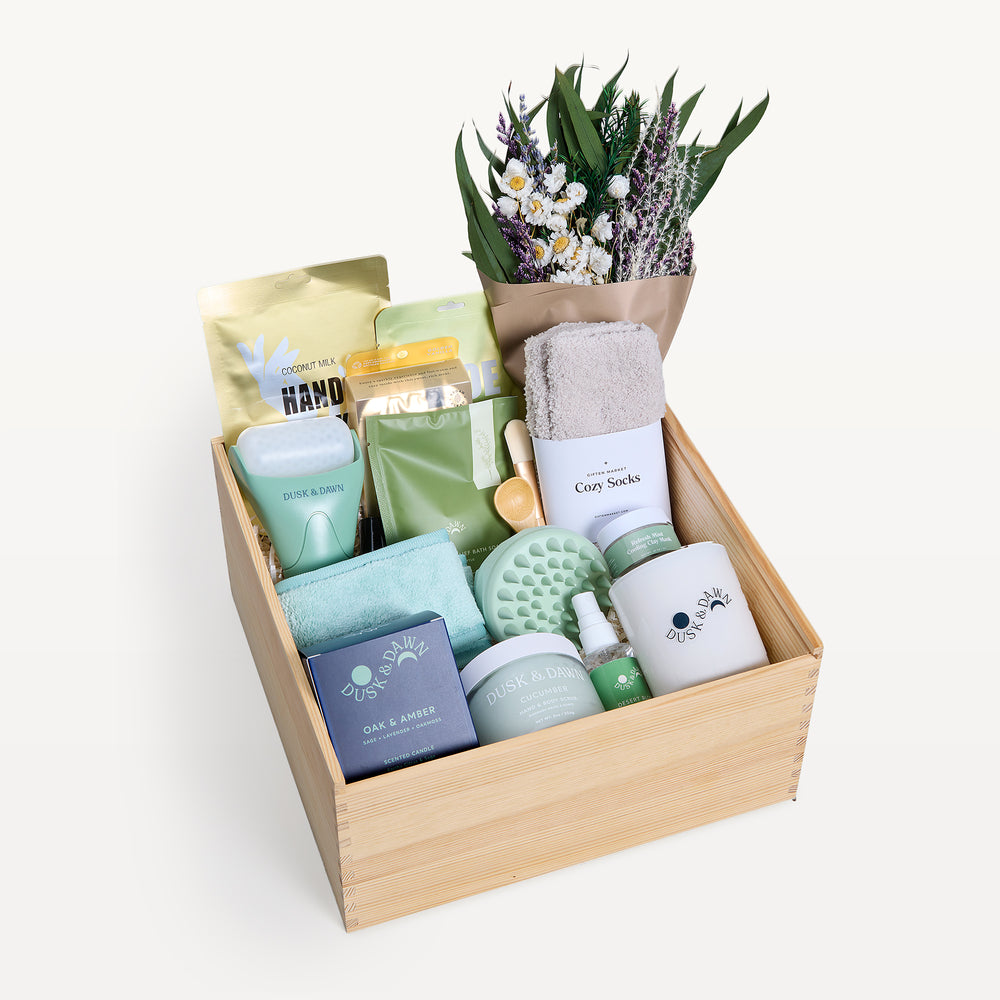 Soothing Sanctuary Deluxe crate open in front, bow-tied lid behind. Dried wildflower bouquet, Cozy Socks, Dusk & Dawn Oak & Amber candle, green scalp massager, coconut milk hand mask, sage-green packets. Giften Market.