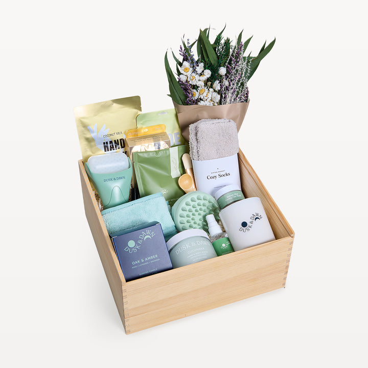 Soothing Sanctuary Deluxe crate open in front, bow-tied lid behind. Dried wildflower bouquet, Cozy Socks, Dusk &amp; Dawn Oak &amp; Amber candle, green scalp massager, coconut milk hand mask, sage-green packets. Giften Market.