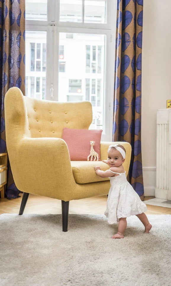 Baby in white dress standing by yellow armchair with giraffe teether in modern living room