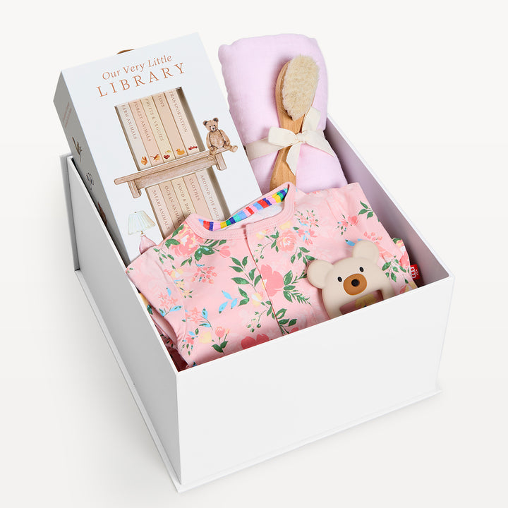 Sweet Beginnings Ainslee, white gift box, lid off. Pink floral zip onesie, Our Very Little Library book set, pink swaddle with bow, wooden brush, bear teether. Giften Market.