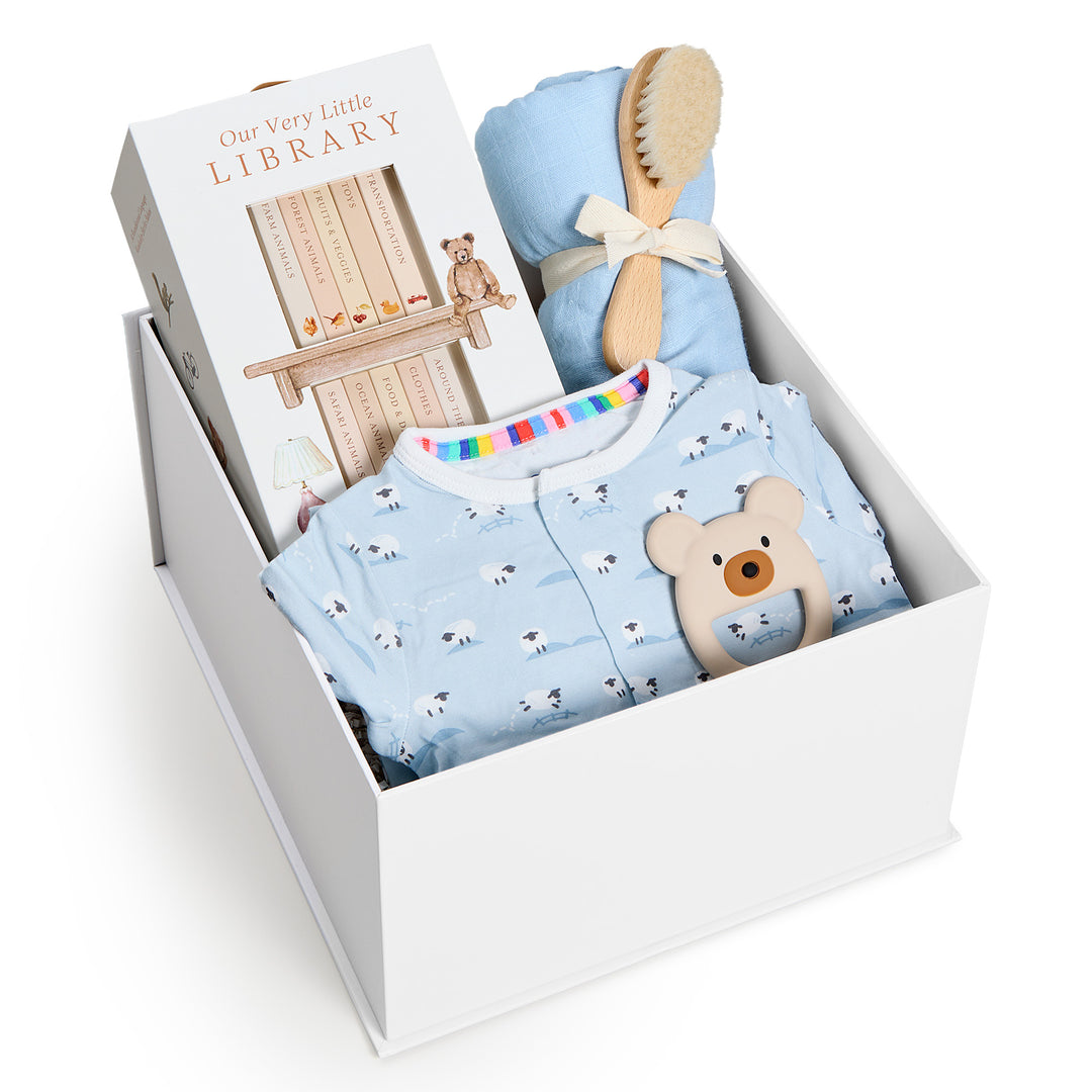 Sweet Beginnings Baa Baa Blue, white gift box, lid off. Blue sheep-print zip onesie, Our Very Little Library, blue swaddle, wooden brush, bear teether. Giften Market.