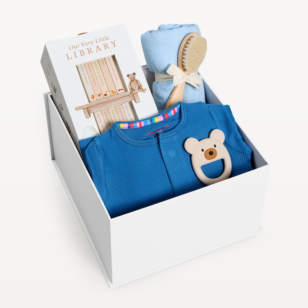 Sweet Beginnings Blue Waffle, white gift box, lid off. Deep blue waffle-knit zip onesie, Our Very Little Library, blue swaddle, wooden brush, bear teether. Giften Market.