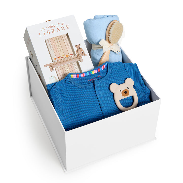 Sweet Beginnings Blue Waffle, white gift box, lid off. Deep blue waffle-knit zip onesie, Our Very Little Library, blue swaddle, wooden brush, bear teether. Giften Market.