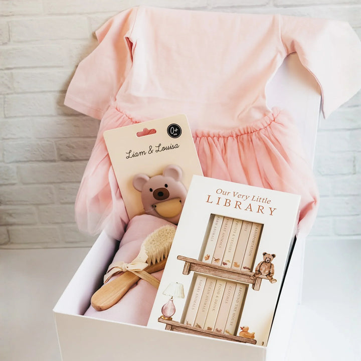 White gift box with pink baby dress, board books, bear pacifier, brush, and blanket