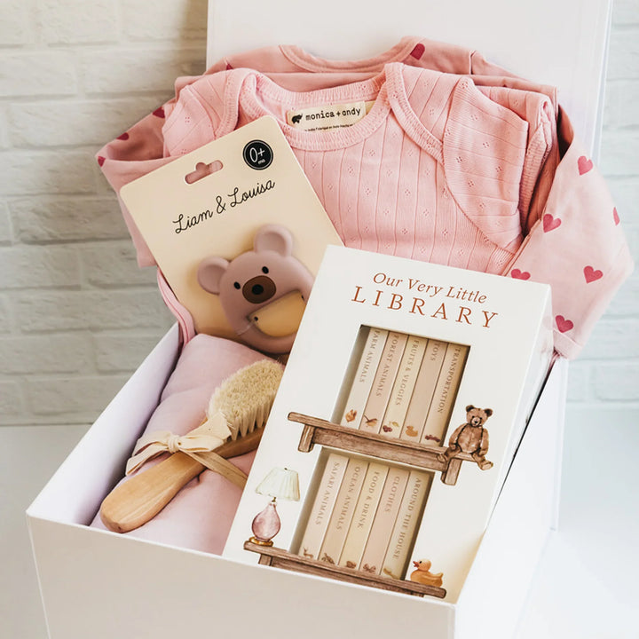 White gift box with pink baby clothes, board books, bear teether, brush, and blanket