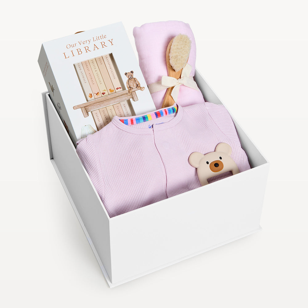 Sweet Beginnings Lilac Snow, white gift box, lid off. Lilac waffle-knit zip onesie, Our Very Little Library, pink swaddle, wooden brush, bear teether. Giften Market.