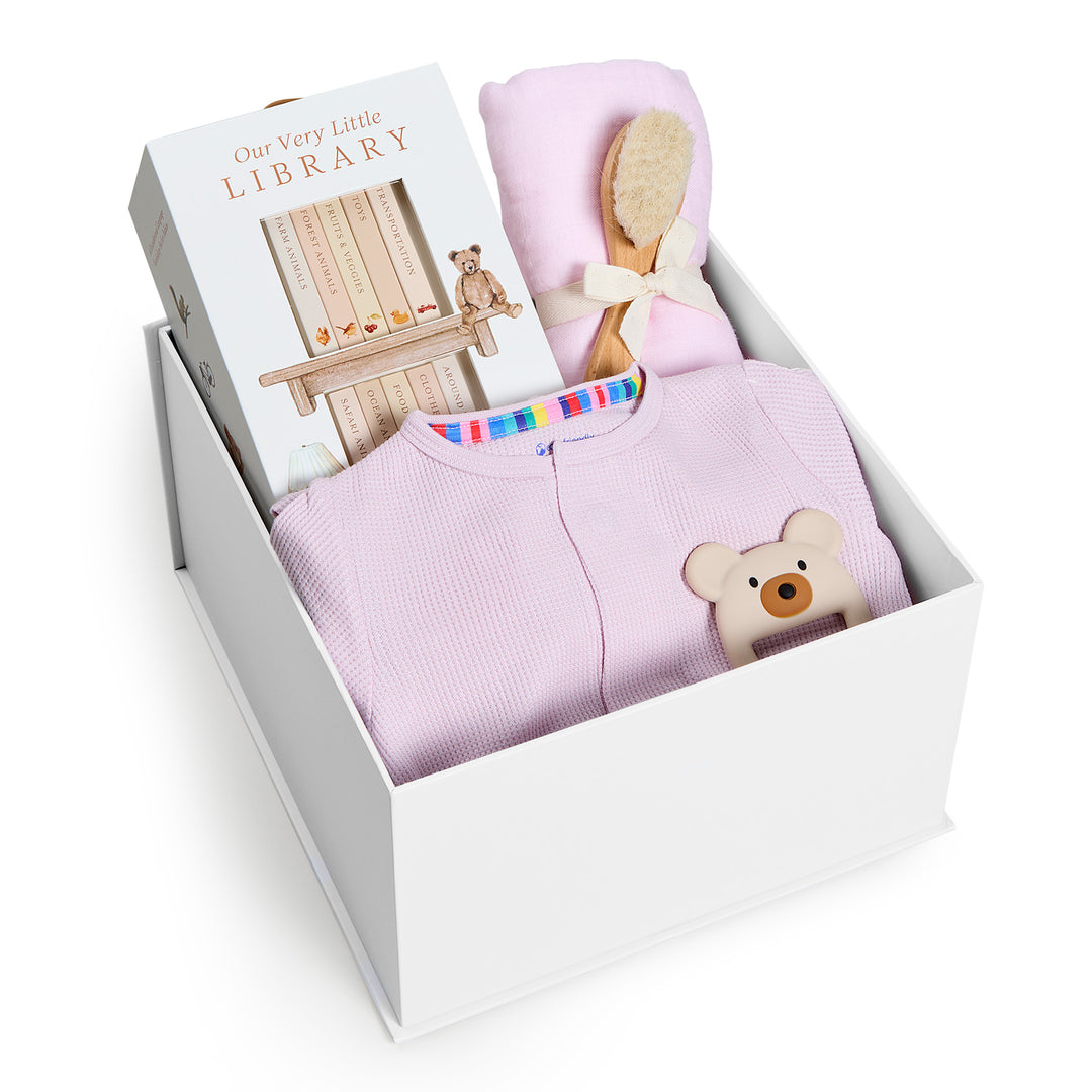 Sweet Beginnings Lilac Snow, white gift box, lid off. Lilac waffle-knit zip onesie, Our Very Little Library, pink swaddle, wooden brush, bear teether. Giften Market.