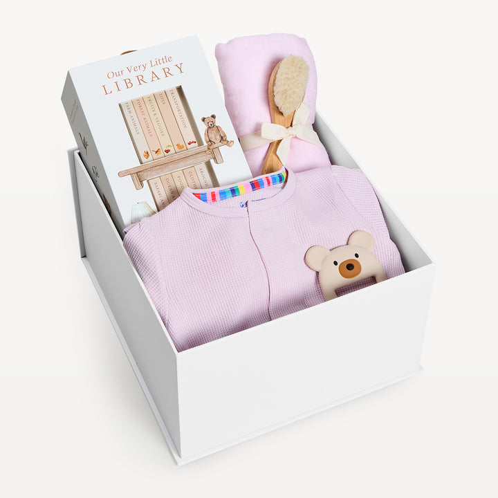 Sweet Beginnings Lilac Snow, white gift box, lid off. Lilac waffle-knit zip onesie, Our Very Little Library, pink swaddle, wooden brush, bear teether. Giften Market.