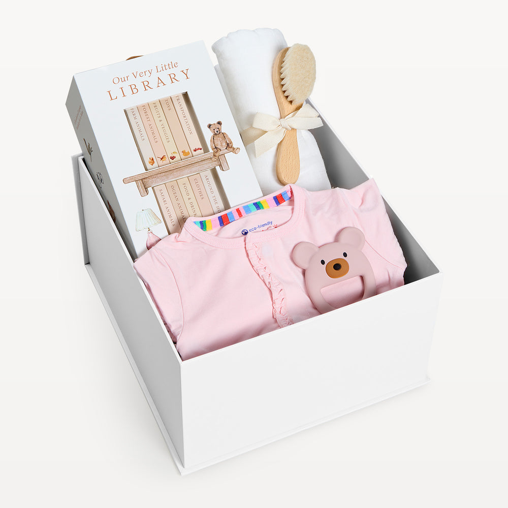 Sweet Beginnings Pink Ruffle, white gift box, lid off. Pink ruffle-shoulder onesie, Our Very Little Library, white swaddle, wooden brush, bear teether. Giften Market.