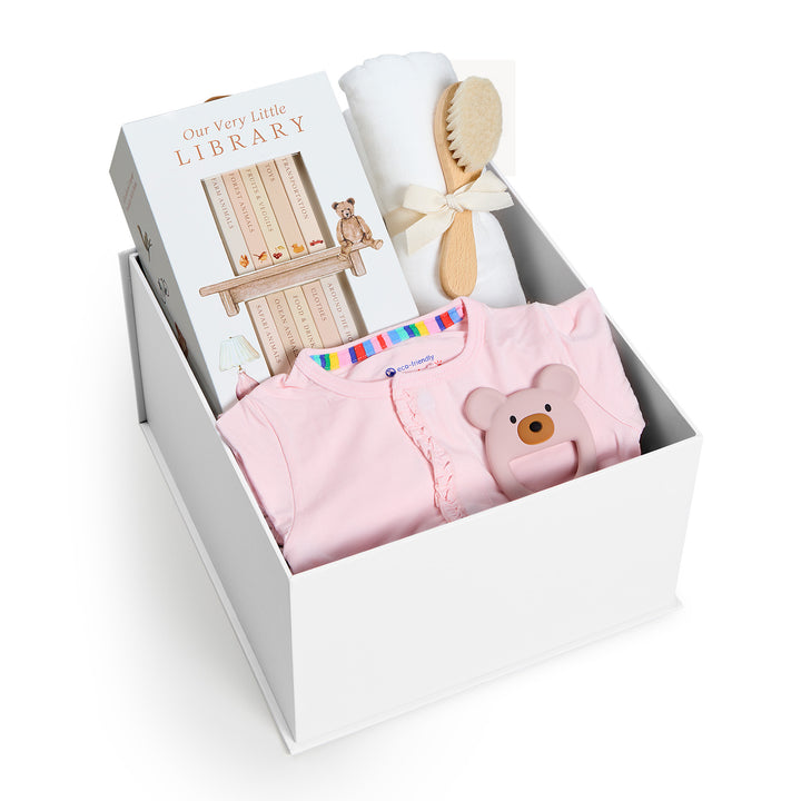 Sweet Beginnings Pink Ruffle, white gift box, lid off. Pink ruffle-shoulder onesie, Our Very Little Library, white swaddle, wooden brush, bear teether. Giften Market.