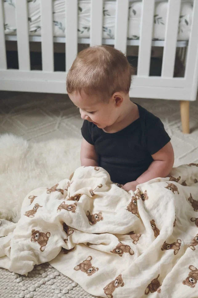 Baby sitting with cream muslin swaddle blanket featuring teddy bear print in nursery setting