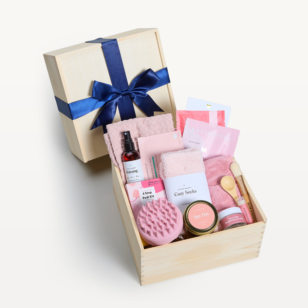 Ultimate Spa Day Classic in a wooden crate, bow-tied lid behind. Pink Cozy Socks, pink scalp massager, Spa Day candle tin, 4 Step Pedi Kit, Relaxing spray, Patchology, waffle hand towel, wooden spoon. Giften Market.