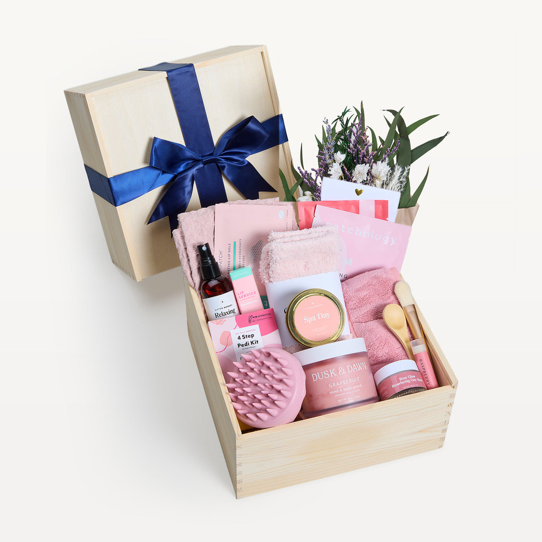 Ultimate Spa Day Deluxe in a wooden crate, bow-tied lid behind, dried bouquet of lavender, white daisies and eucalyptus. Pink Cozy Socks, scalp massager, Spa Day tin, Pedi Kit, Dusk & Dawn Grapefruit body butter, spray, towel, wooden spoon. Giften Market.