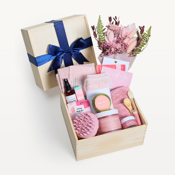 Ultimate Spa Day Deluxe in a wooden crate, bow-tied lid behind, pink dried bouquet. Pink Cozy Socks, scalp massager, Spa Day tin, Pedi Kit, Dusk &amp; Dawn Grapefruit body butter, spray, towel, wooden spoon. Giften Market.