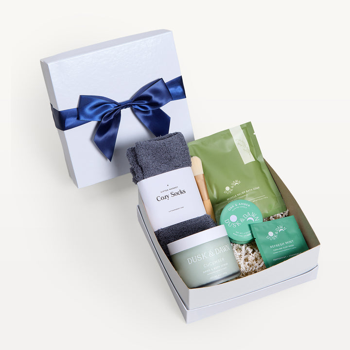 Wellness Ritual, white shallow box open in front, bow-tied lid behind. Charcoal Cozy Socks, green pouch, Dusk &amp; Dawn Cucumber jar, Refresh Mint clay mask, small tin, wooden spoon. Giften Market.